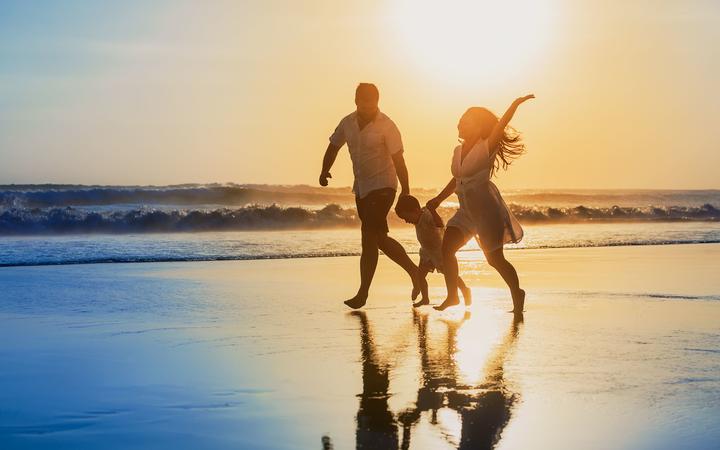 Happy family - father, mother, baby son hold hands and run with fun along edge of sunset sea on black sand beach. Active parents and people outdoor activity on tropical summer vacations with children
