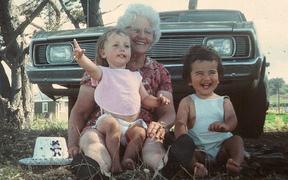 Two babies on knee of an older woman on the grass in front of a 70s car.
