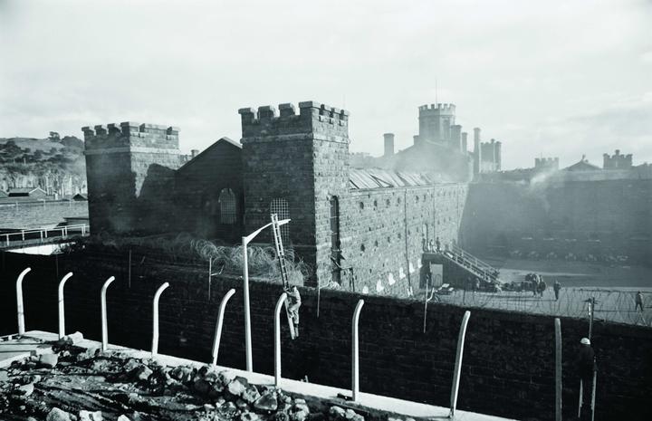 Mount Eden Prison on fire during rioting in 1965. In the foreground, Auckland Electric Power Board staff erect floodlights. 