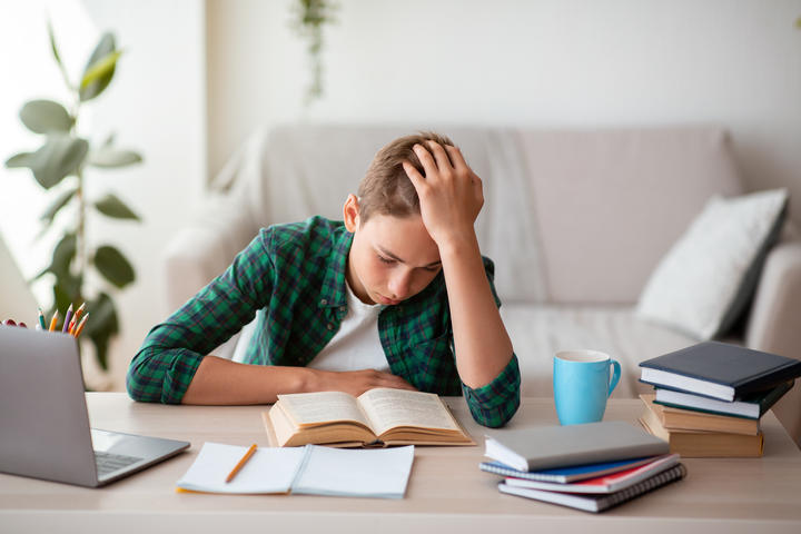 Bored teenager reading book at desk at home, holding his head, free space