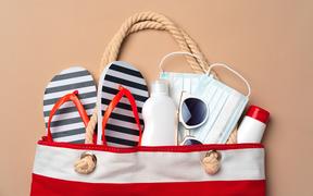 Beach bag with beach items and medical protective mask on beige background. Coronavirus summer concept