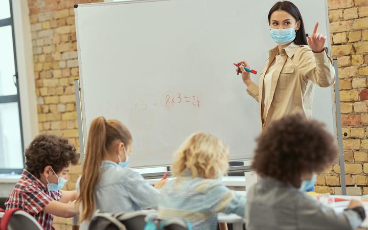 在冠状病毒大流行期间戴着防护面具的女教师在船上为一群小学生写数学任务。 教育、数学和 covid19 概念