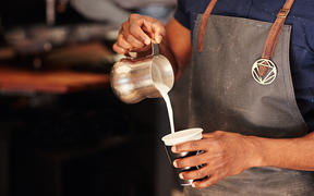 Cropped image of an African barista carefully pouring milk from a stainless steel jug into a takeaway cup in a coffee shop