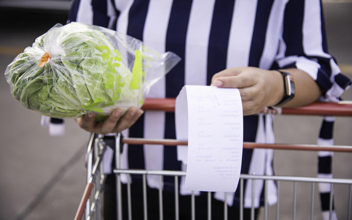 Woman shopping with trolley, holding receipt of grocery items, with food in handing, checking prices.