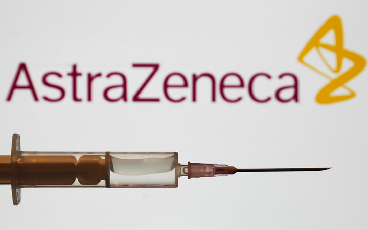 Medical syringe is seen with AstraZeneca company logo displayed on a screen in the background.