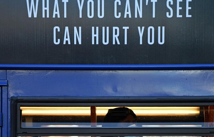 A woman wearing a face mask or covering due to the COVID-19 pandemic, sits beneath a sign reading "What you can't see, Can hurt you", as she travels by bus in Manchester.