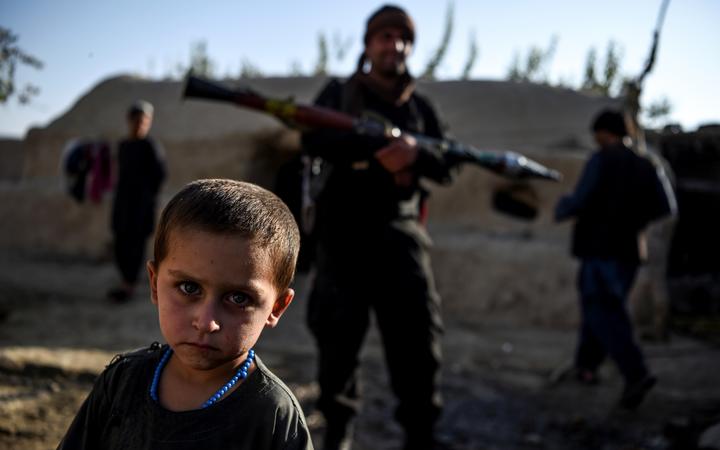 A young boy looks at the camera as a policeman holding a rocket-propelled grenade stands behind in a house at Deh Qubad village in Maiwand district of Kandahar province on September 27, 2020. 