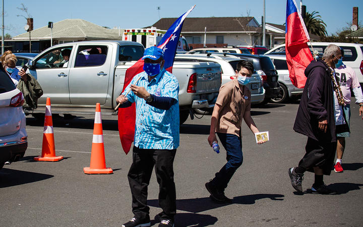 A man waving a Samoan flag and blue face covering dances at the Browns Road netball courts in Manurewa.