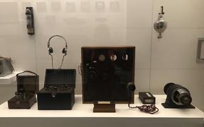 Shows a display case with some early radio equipment used by Professor Robert Jack. A large wooden framed transmitter sits in the centre with a generator and microphone to the side.