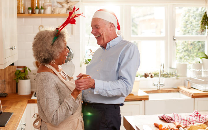 Loving Senior Couple Wearing Fancy Dress Antlers Dance In Kitchen Whilst Preparing Christmas Dinner