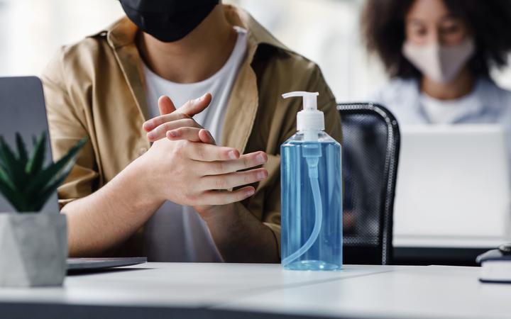 Focus on antiseptic to protect against covid virus. Guy disinfects his hands, sitting at table at workplace with laptop, working in interior of modern office, panorama, copy space