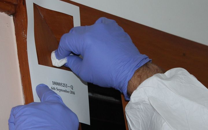 A close up photo of two hands with gloves on, swabbing within a 100 square centimetre stencil stuck to a wall. By swabbing within that area, testing companies get a sample that corresponds to Ministry of Health guidelines for meth. 