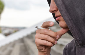 A teenage boy wearing a hoodie smokes a cigarette.