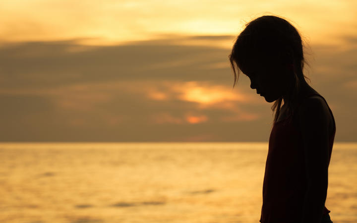 Portrait of sad blond little girl standing on the beach at sunset time.