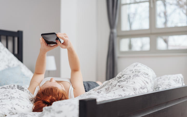 Young woman spending a relaxing day at home lying on her back on the bed reading a message on her mobile phone